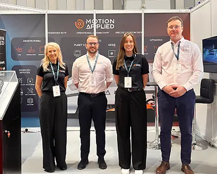 Four people standing side by side at an exhibition stand with a backdrop displaying the “Motion Applied” logo and product sections. Each person wears a lanyard badge. The stand includes panels with headings such as “Architectures,” “Management Tools,” “Applications,” and “Networks,” along with a counter, chairs, and equipment in the background.