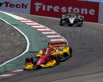 Two INDYCARs race down the corkscrew at Laguna Seca Raceway, with a red and yellow car leading and another car following.
